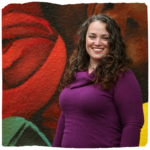 Dena Zaldua smiles brightly in a regal purple sweater in front of a richly-hued mural in Southeast Portland.