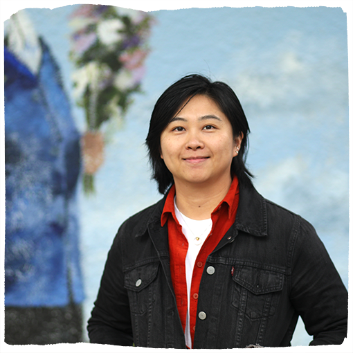 Sandee Huang poses in front of a light blue mural in Southeast Portland wearing a black denim jacket and a red collared shirt.