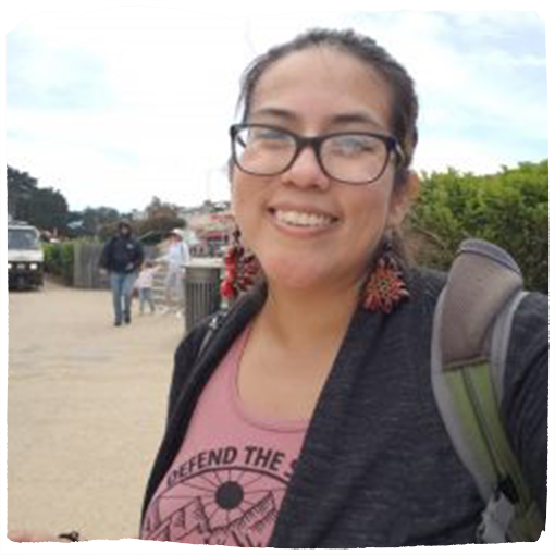 Ana Molina wears a dusty rose colored shirt and a backpack while smiling out in the field.