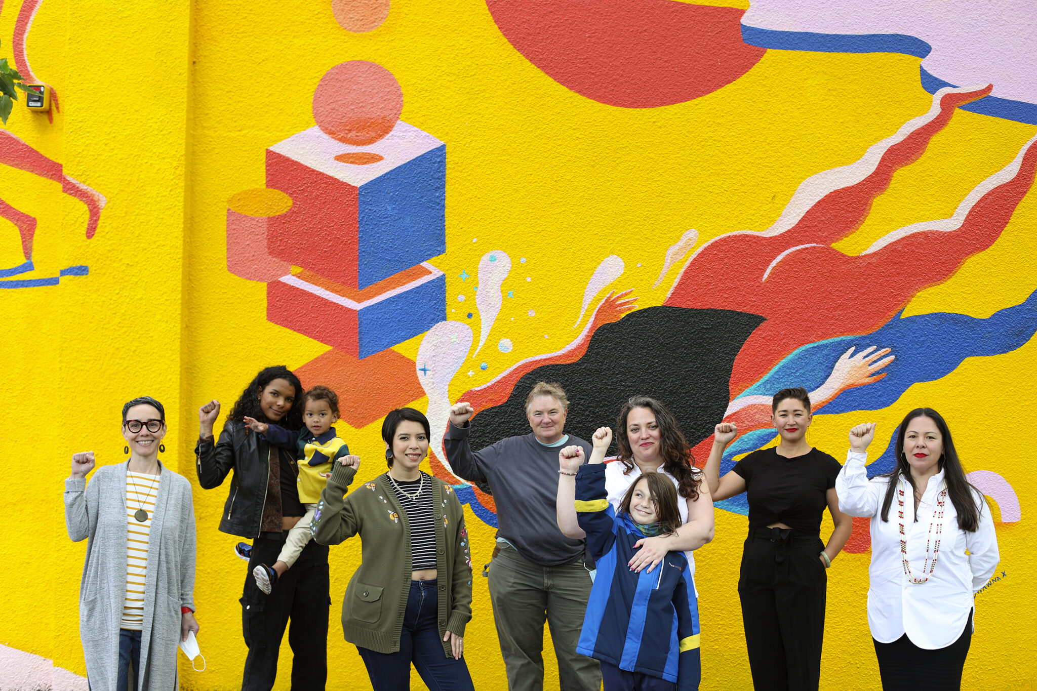 The Seeding Justice staff stand in front of a bright yellow mural with their fists raised up.