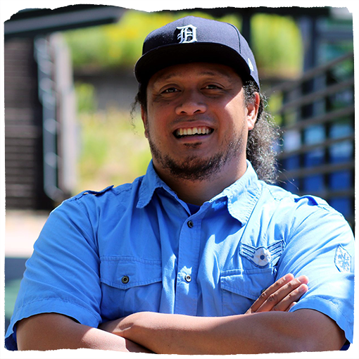 Makerusa smiles with his arms crossed outside, wearing a light blue, short-sleeved shirt and a baseball cap in the sun.