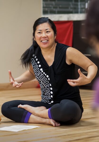 Michelle Fujii sits cross-legged on the floor, hands raised chest height and smiling as if getting prepapred.