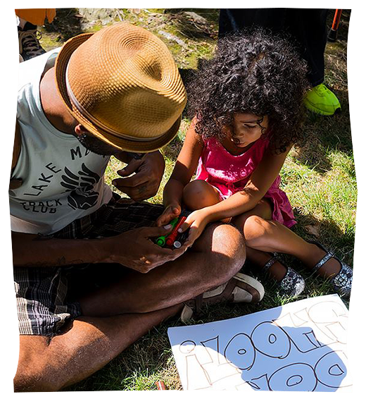 A person and a young child draw a protest sign together while sitting cross-legged on the grass.