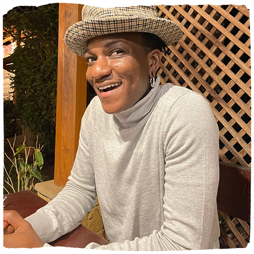 Silver grins wearing a light gray turtleneck and printed fedora in front of a wooden lattice fence.