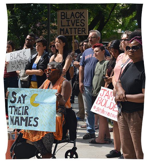 A crowd holding protest signs with "say their names" and "black lives matter" stand quietly while listening to a speaker, out of frame.