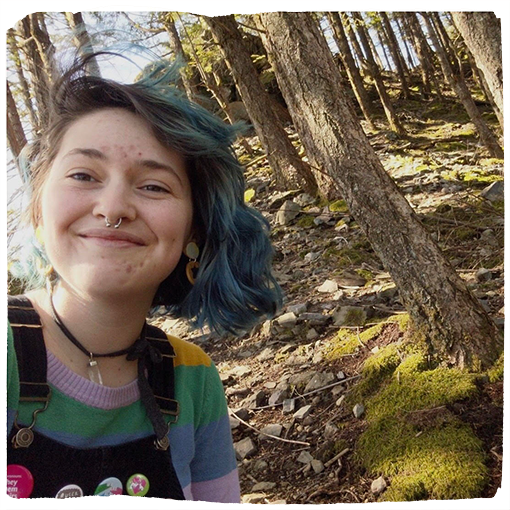 Davis Esther Rose wears a rainbow colored long sleeve shirt under black overalls adorned with pins. They're smiling into the camera with blue hair in front of a grove of trees.