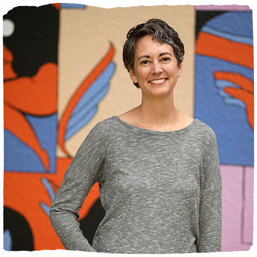 Wendy Cluse wears a gray, long sleeve shirt while smiling in front of a pale yellow and blue mural in Southeast Portland.