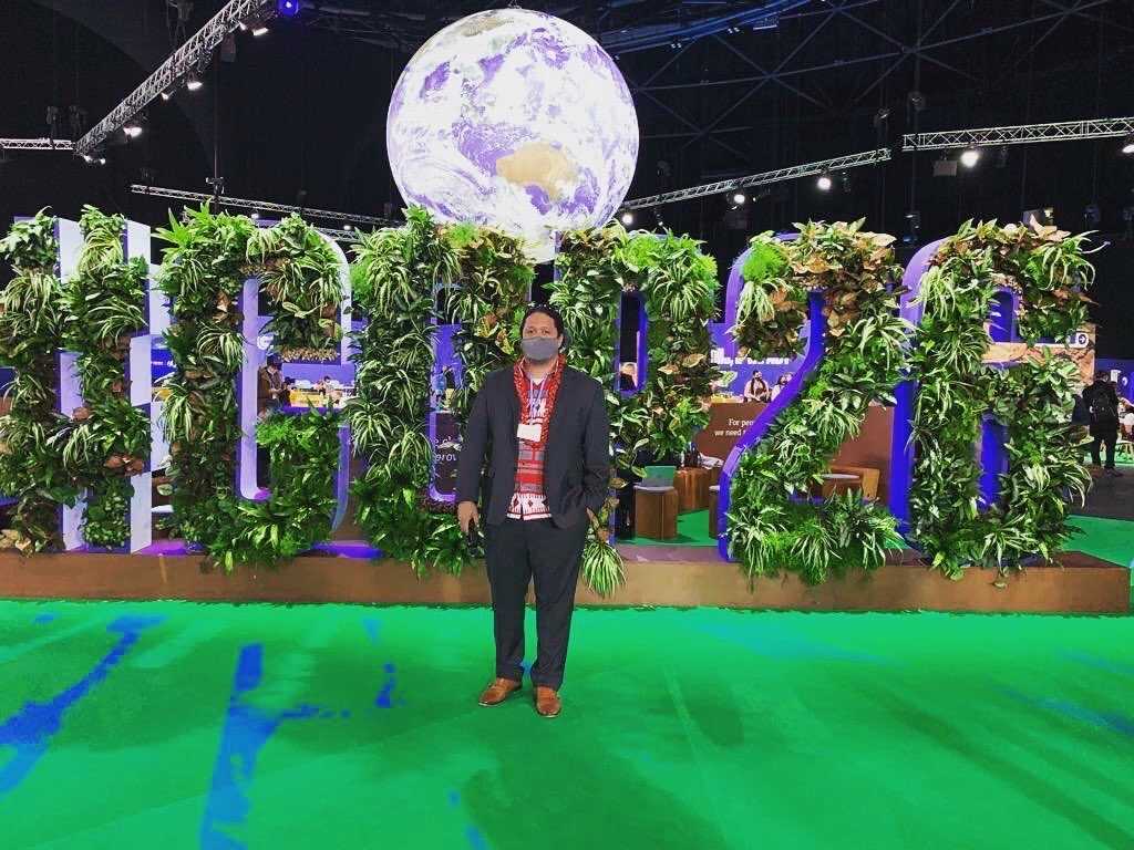 Makerua Porotesano stands in front of a life-size, plant-covered, COP26 cut-out.