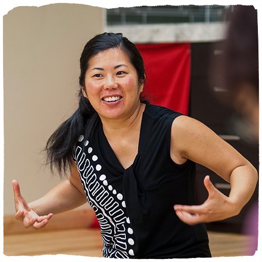 Taiko artist Michelle Fujii sits cross-legged on the floor with her arms raised.