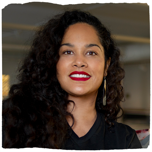 Wearing deep red lipstick and dark curly hair, Natalie Ball smiles at the camera.