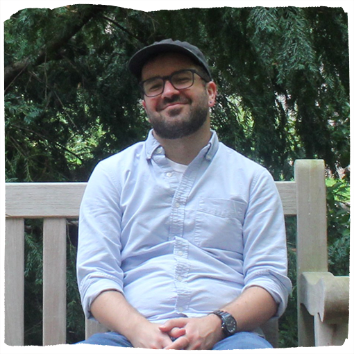 Ryan smiles while sitting on a bench in a blue baseball cap and a light blue button down .