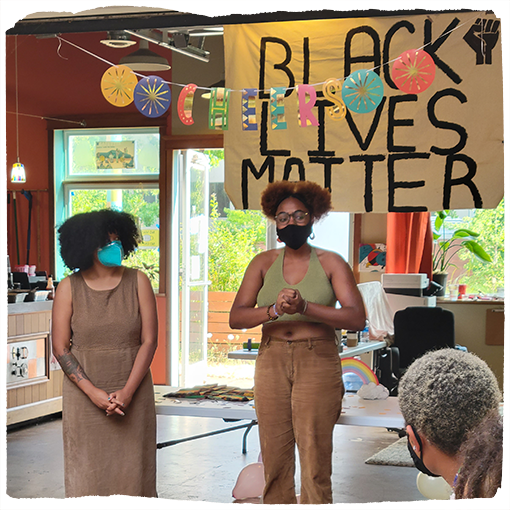 Two people wearing masks speak to a group in front of a Black Lives Matter sign.